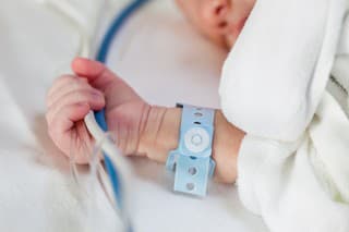 Close-up of a caucasian newborn baby holding life-support hoses and cables tight in his had with wrist nametag while sleeping in a hospital bed.