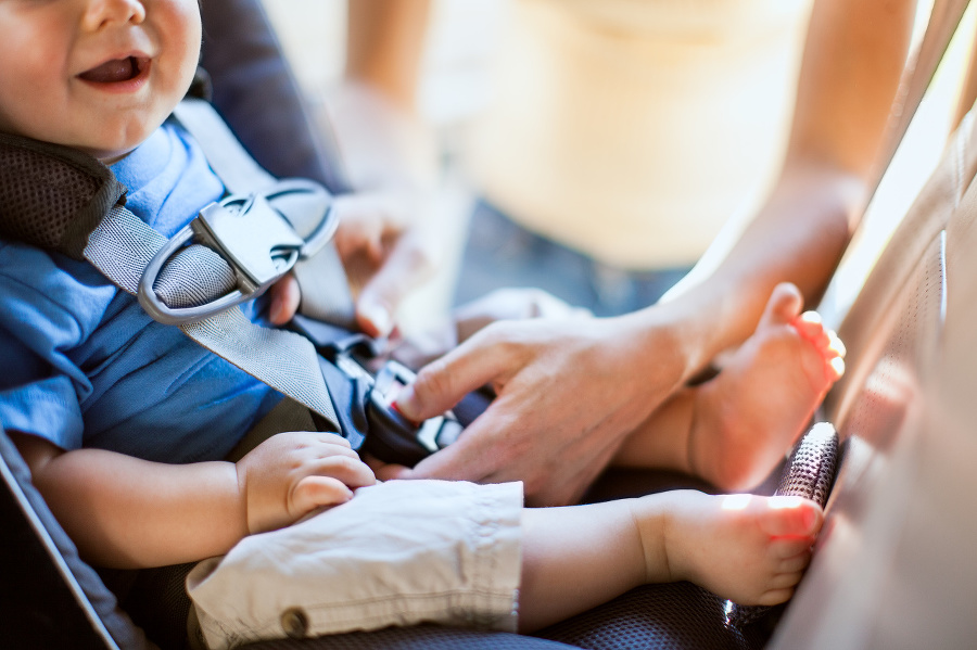 A mom buckles her infant son safely into a rear facing car seat as they get ready to drive somewhere in their vehicle.  The baby's mouth is visible with a big smile.  Sunlight shines in from behind.  Horizontal with copy space.