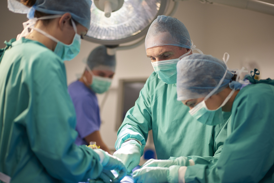 a female operating nurse stands over a patient at the operating table and looks down to what she is doing in the operation. She is joined by a young female nurse and mature male surgeon .In the background the anaesthetist is looking over from his monitors