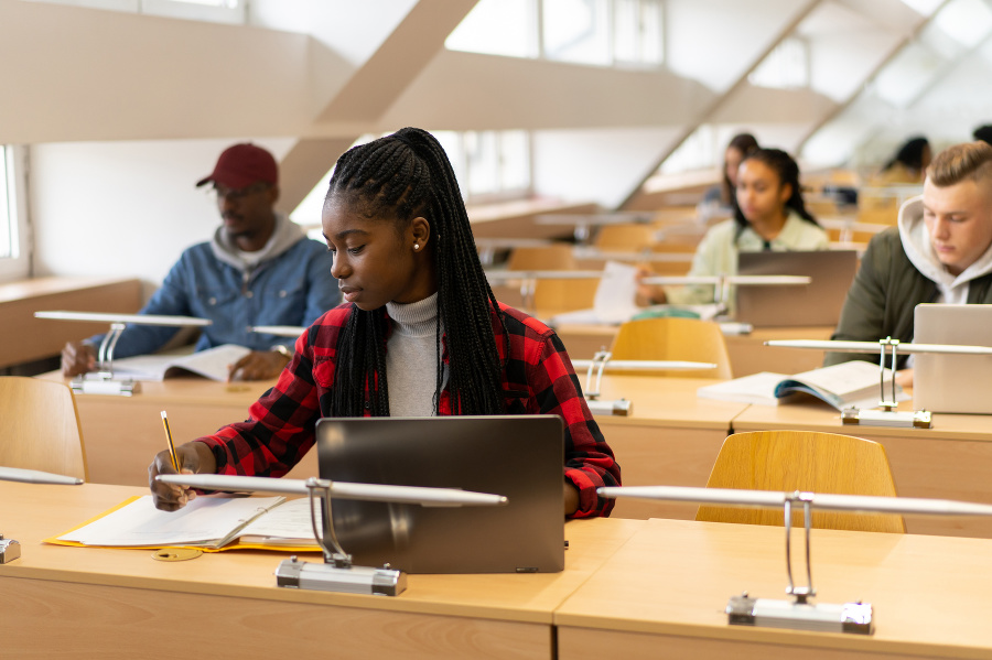 Young afro-American student is writing in the library. More students are sitting behind her
