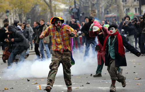 Aktivisti v maskách klaunov utekajú pred políciou medzi vybuchujúcimi granátmi slzného plynu počas demonštrácie za lepšie životné prostredie v Paríži.