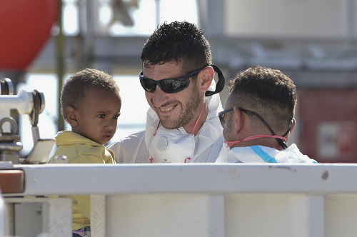 Dieťa neveriacky pozerá na usmiatych príslušníkov pobrežnej stráže v prístave Messina. Do Talianska mieria denne tisícky ľudí cez Stredozemné more.