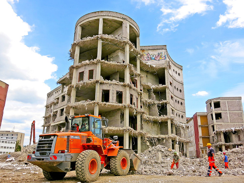 Stavbu postupne obhrýza demolačné pásové rýpadlo.