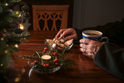 Hands of an elderly single man sitting alone at a table with Christmas cookies, coffee and festive decoration next to an empty chair, lonely holidays during the croronavirus pandemic or after a loss, selected focus