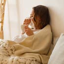 Sick young woman lying in bed covered with blanket holding a cup of hot tea